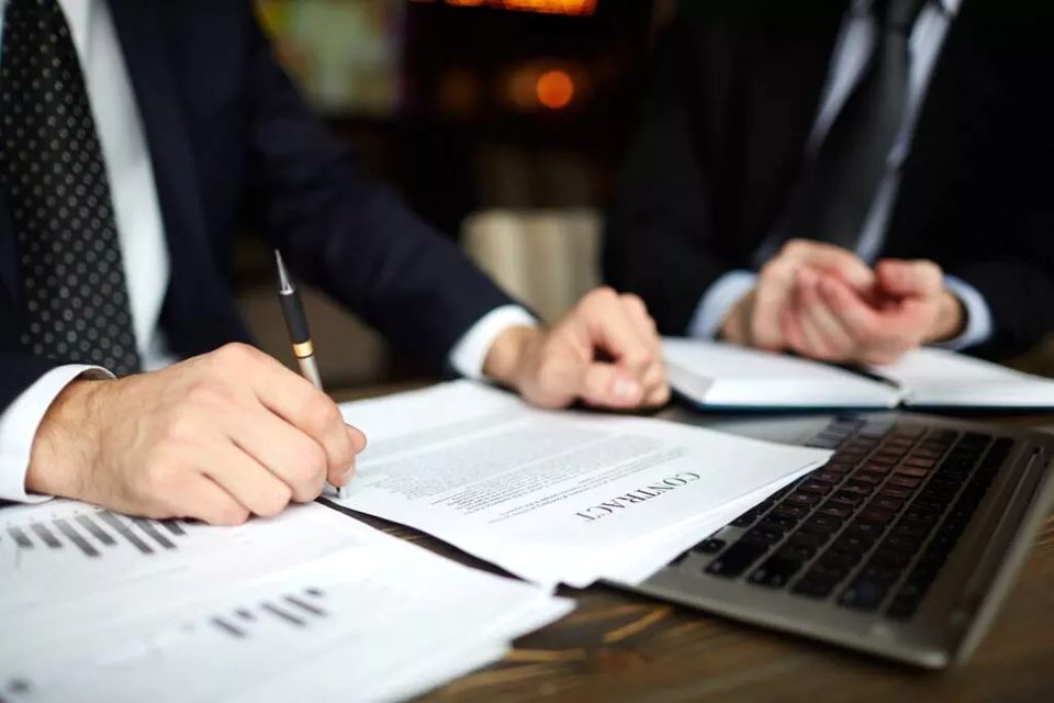 Assinatura de documento sobre a mesa, ao lado de relatórios e notebook